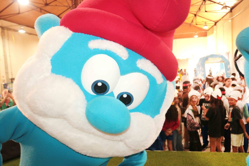 Personagem dos Smurfs em tamanho gigante interage com o público em um espaço repleto de estudantes participantes do evento.