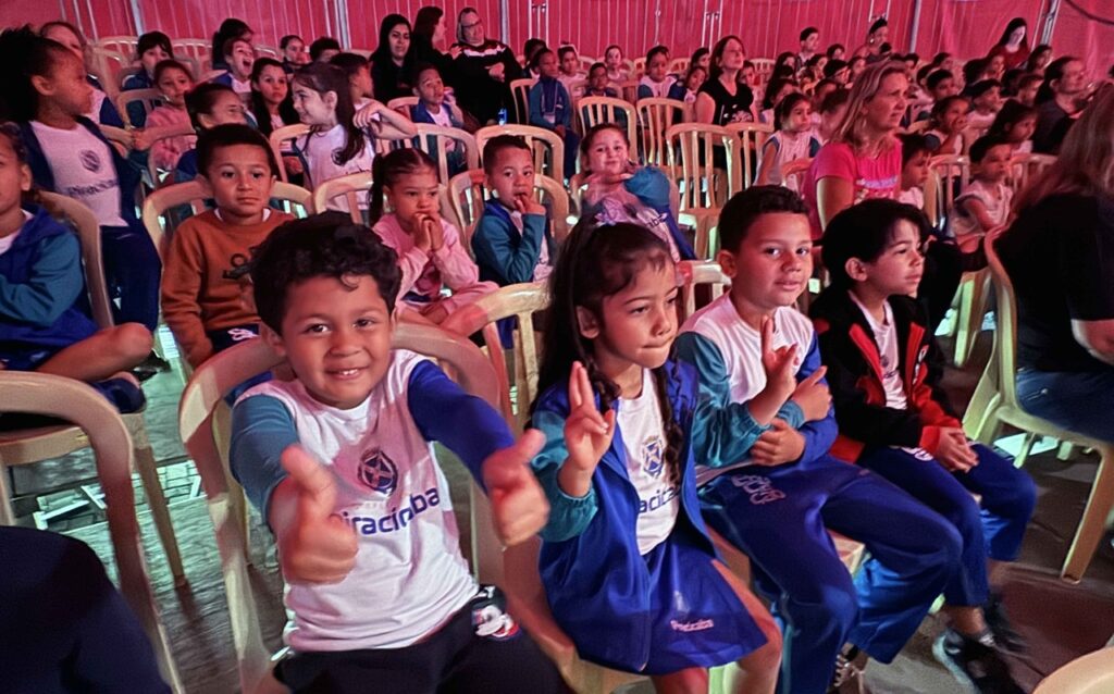 A imagem mostra uma plateia formada principalmente por crianças, sentadas em cadeiras plásticas dentro de uma tenda ou espaço de evento com iluminação colorida. Em primeiro plano, algumas crianças usam uniformes com o nome “Piracicaba”; um menino faz sinal de positivo para a câmera, enquanto outras crianças fazem gestos e sorriem. Ao fundo, há muitas crianças e adultos acompanhando o evento. As paredes são vermelhas e o ambiente está bastante cheio, com luzes verdes e rosadas refletindo no chão e nas cadeiras.
