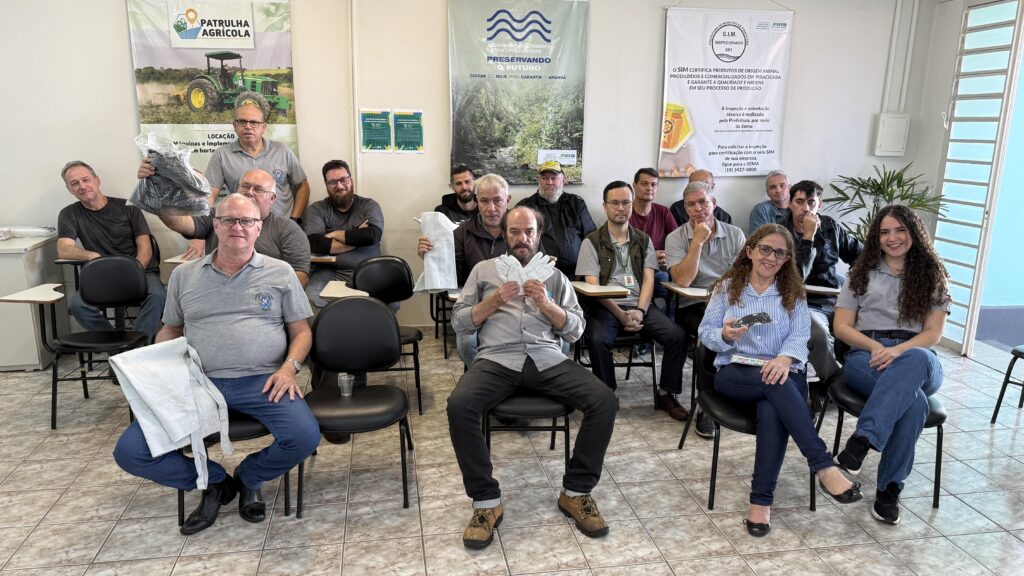 A imagem mostra um grupo de participantes de treinamento reunidos em uma sala. São cerca de quinze pessoas sentadas em cadeiras, algumas segurando materiais de trabalho como aventais ou uniformes. O ambiente é simples, com piso de cerâmica e paredes claras, decoradas com cartazes institucionais sobre programas agrícolas e ambientais — como “Patrulha Agrícola” e “Preservando o Futuro”.