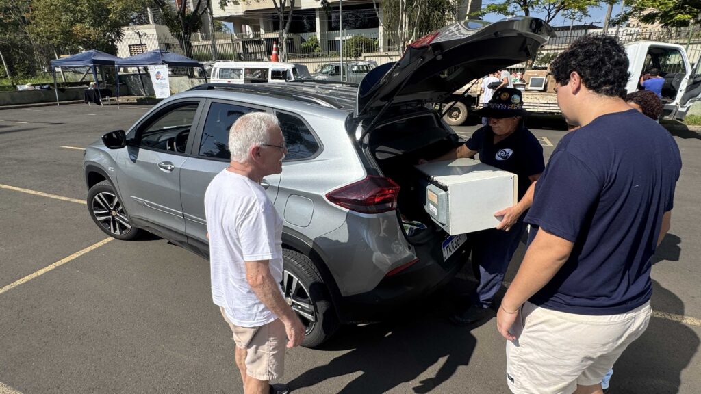 Pessoas carregam uma caixa com equipamentos eletrônicos para o porta-malas de um carro.
