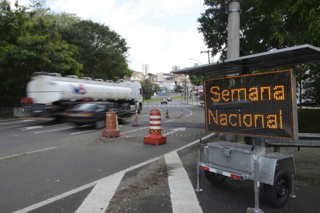A imagem mostra uma via urbana com tráfego de veículos e um painel eletrônico móvel instalado na lateral da rua. No painel iluminado aparece a mensagem “Semana Nacional”, provavelmente usada para orientar ou informar motoristas.
