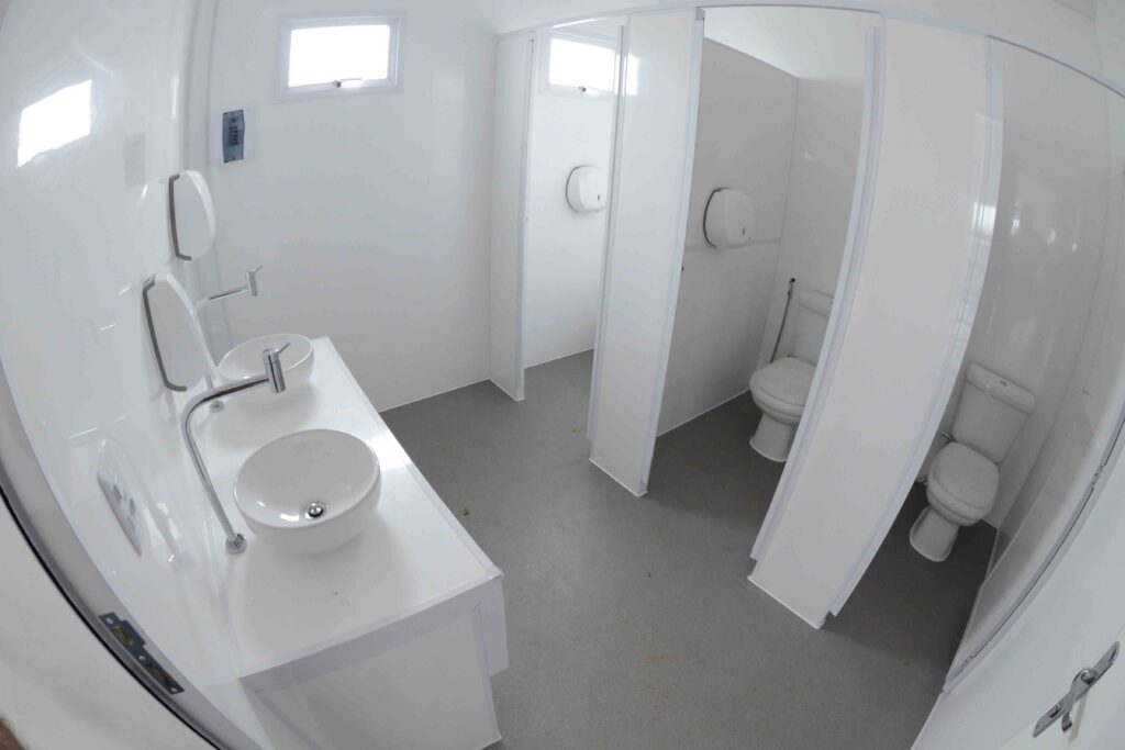 Esta fotografia em grande angular mostra o interior de um banheiro público moderno e limpo, destacando pias brancas à esquerda e baias individuais com vasos sanitários à direita, criando uma atmosfera organizada e funcional. A composição é capturada a partir de um ponto de vista elevado e amplo, utilizando uma lente que distorce ligeiramente as linhas para enfatizar o espaço aberto. À esquerda, uma bancada longa e branca acomoda duas pias redondas de sobrepor com torneiras cromadas altas. O piso cinza escuro estende-se por todo o ambiente, conectando a área das pias às cabines dos banheiros localizadas do lado direito. As divisórias brancas separam claramente cada compartimento, guiando o olhar do observador em direção aos fundos da sala. Os principais elementos em destaque são as duas pias circulares brancas, elegantes e minimalistas, equipadas com torneiras de metal brilhante. A bancada onde estão instaladas possui acabamento liso e brilhante, refletindo a iluminação ambiente. Mais ao fundo, nas cabines individuais, aparecem dois vasos sanitários brancos de louça, limpos e bem posicionados, acompanhados por dispensadores de papel higienizado fixados nas paredes divisórias. A imagem é uma fotografia digital com uma estética limpa, típica de portfólios arquitetônicos ou de design de interiores. A execução técnica utiliza uma lente grande angular para maximizar a percepção de espaço em uma área possivelmente compacta. A paleta de cores é predominantemente neutra, baseada em tons de branco, cinza e prata, o que transmite uma forte sensação de higiene, ordem e modernidade. O ambiente é um banheiro interno e fechado, iluminado uniformemente por luzes artificiais que se refletem nas superfícies brilhantes das paredes e bancadas. Pequenas janelas no alto das paredes permitem a entrada de luz natural suave, complementando a iluminação geral. A atmosfera geral é de extrema limpeza, funcionalidade e sobriedade, típica de instalações comerciais ou públicas bem cuidadas.