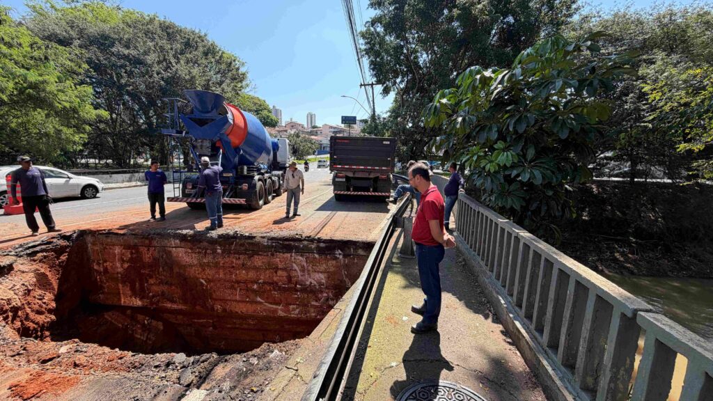Equipe realiza obra de manutenção viária em ponte, com escavação de grande porte e caminhões de apoio.