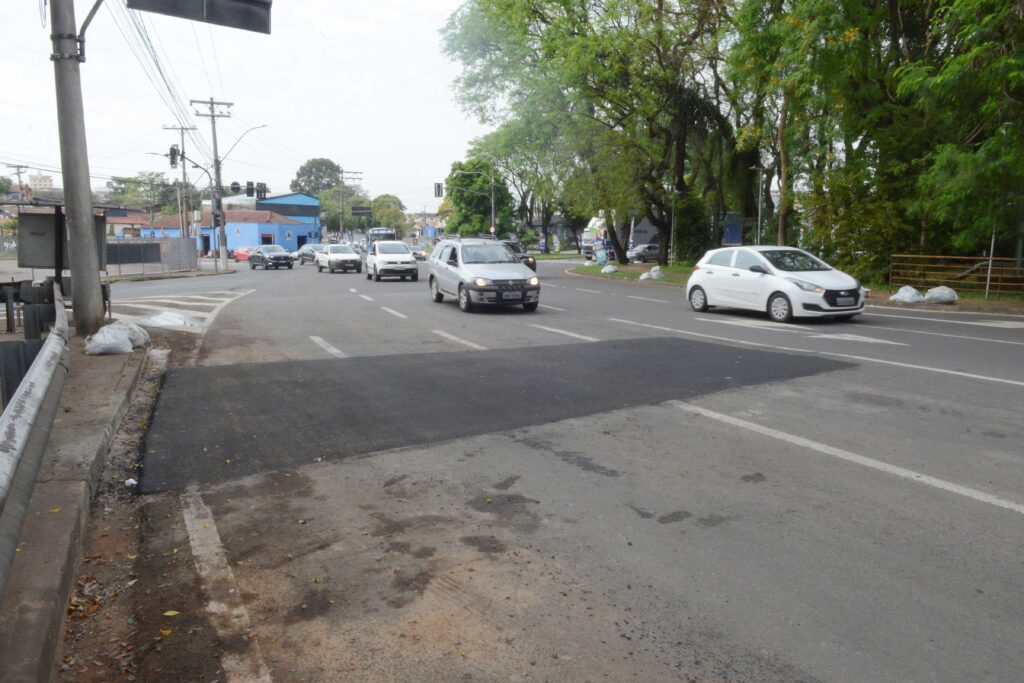 Perspectiva oposta da mesma via, mostrando a faixa recente de asfalto recém-aplicado no pavimento, com tráfego contínuo de veículos e área arborizada ao lado.