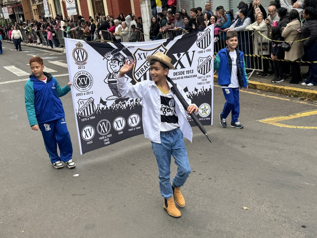 Na imagem, vemos um desfile festivo em Piracicaba, onde três crianças caminham pela rua segurando um grande banner do Esporte Clube XV de Novembro de Piracicaba, destacando sua história desde 1913 com emblemas de diferentes épocas e o mascote animado do clube erguendo um troféu, cercado por torcedores; o ambiente é alegre e comunitário, com espectadores atrás de grades acompanhando a celebração que exalta o orgulho local pelo tradicional time de futebol.