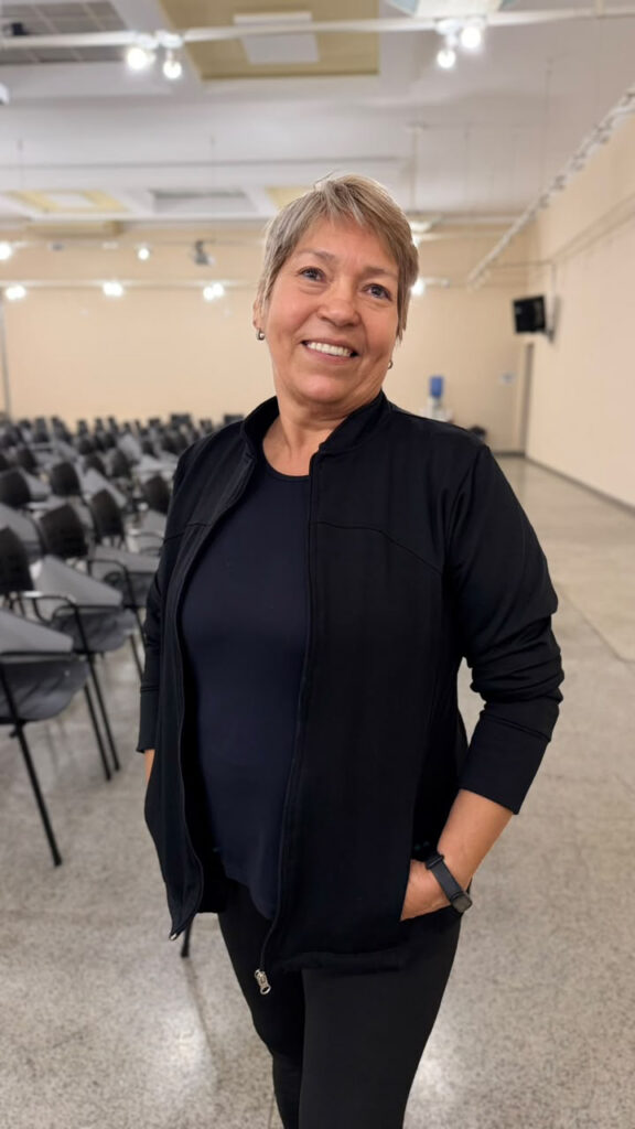 A imagem mostra uma mulher sorridente em um ambiente interno amplo, com várias cadeiras organizadas em fileiras — provavelmente um auditório ou sala de eventos.
