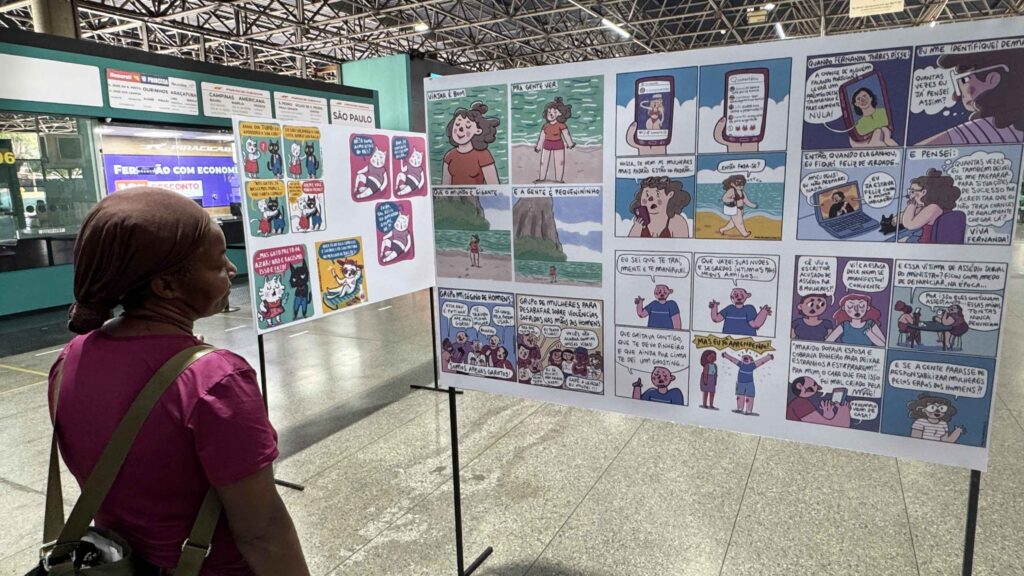 A imagem mostra uma mulher, vista de costas, observando uma exposição de histórias em quadrinhos dentro de um terminal rodoviário. Ela usa uma blusa vinho e carrega uma bolsa transversal. Os painéis apresentam ilustrações coloridas e textos em português, abordando temas como relacionamentos, machismo, violência contra a mulher e situações do cotidiano. Ao fundo, aparecem guichês e placas de destinos, incluindo Campinas, Americana e São Paulo, além de um painel luminoso com a mensagem “Feriadão com economia”.