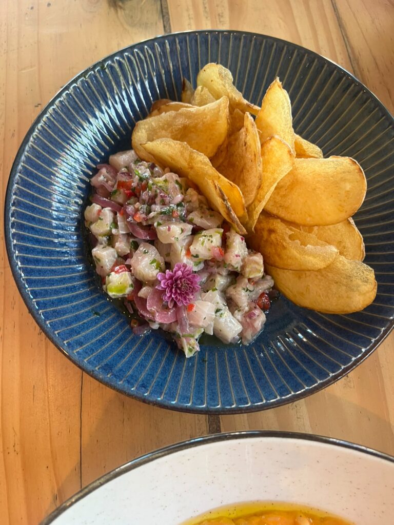 O prato apresenta uma composição elegante e colorida: um ceviche de peixe branco cortado em cubos, misturado com cebola roxa, pimentas e ervas frescas, decorado com uma flor comestível roxa no topo. Ao lado, há chips de batata dourados e crocantes, servidos em um prato azul com textura radial que realça as cores dos ingredientes.