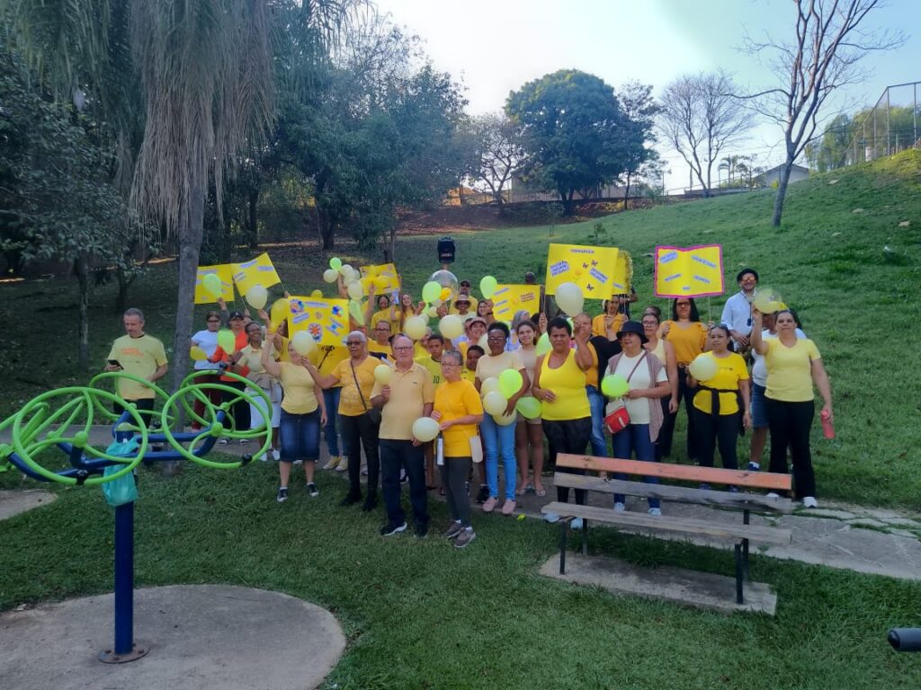 Uma vibrante fotografia ao ar livre retrata um grande grupo de pessoas reunidas em um parque arborizado, vestidas predominantemente com camisas amarelas e segurando balões e cartazes, transmitindo uma atmosfera de conscientização e união comunitária. A imagem apresenta uma composição aberta e dinâmica, com dezenas de participantes dispostos em várias fileiras em uma encosta gramada, criando profundidade visual e engajamento. Algumas pessoas estão posicionadas mais à frente, próximas a um banco de madeira e a equipamentos de ginástica verde-claros ao ar livre, enquanto outras se espalham pelo talude e ao fundo. A ação central captura um momento estático e posado, onde os indivíduos olham diretamente para a câmera, erguendo cartazes amarelos informativos, balões coloridos e sorrindo, demonstrando coesão e propósito coletivo. Os participantes são de diversas idades, etnias e gêneros, exibindo uma ampla variedade de feições, expressões alegres e posturas acolhedoras. A maioria veste camisas ou blusas em tons de amarelo vibrante, combinadas com calças jeans, escuras ou bermudas, criando uma forte identidade visual unificada. As pessoas nas primeiras fileiras destacam-se com clareza, exibindo sorrisos genuínos e segurando firmemente balões amarelos e verdes, além de cartazes decorados com ilustrações e textos voltados para a conscientização. Trata-se de uma fotografia digital realista, caracterizada por uma captura clara e nítida sob luz natural diurna, com foco acentuado nos sujeitos em primeiro e segundo plano. A paleta de cores é dominada por tons terrosos da grama e do solo, o verde exuberante da vegetação circundante e o amarelo marcante das roupas, balões e cartazes, que servem como ponto focal cromático. A execução técnica prioriza a nitidez e o registro documental, equilibrando a exposição entre as áreas ensolaradas e sombreadas da cena. A cena se desenrola em um parque público bem cuidado, caracterizado por um gramado verde, árvores frondosas, arbustos e uma encosta suave que eleva o plano de fundo. A iluminação é difusa e natural, vinda de um dia parcialmente ensolarado, projetando sombras suaves e criando um ambiente acolhedor e comunitário. O cenário ao ar livre, combinado com a energia positiva dos participantes, reforça um clima de solidariedade, engajamento social e esperança.