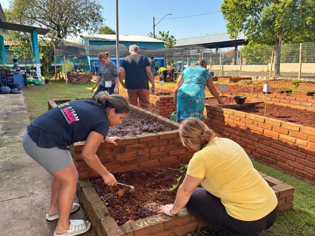 Grupo de pessoas realiza atividades de jardinagem em canteiros elevados de uma horta comunitária. Os participantes estão preparando a terra e cuidando das plantas em um espaço ao ar livre, cercado por áreas verdes e estruturas de apoio. A cena destaca uma ação coletiva de cultivo e manutenção dos canteiros.