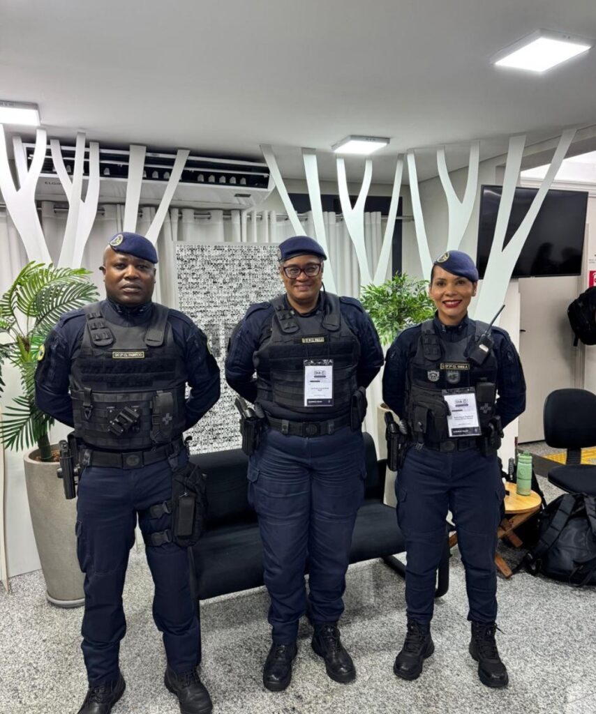 A imagem mostra três guardas civis municipais uniformizados, posando lado a lado em um ambiente interno moderno. Eles vestem uniformes azul-escuros com coletes táticos e boinas, transmitindo uma postura profissional e de representação institucional.