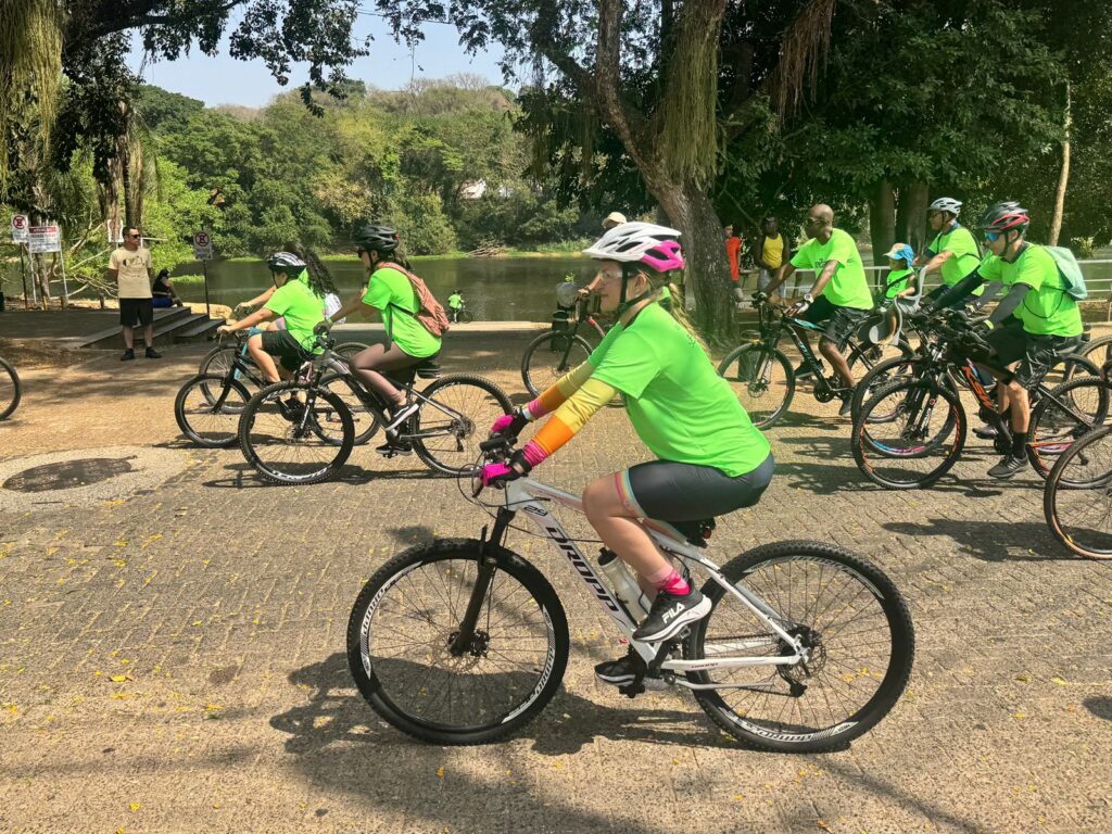 A imagem mostra um grupo de ciclistas participando de um passeio ou evento ao ar livre. Todos usam camisetas verdes, capacetes de segurança e estão em bicicletas de diferentes modelos, principalmente mountain bikes.