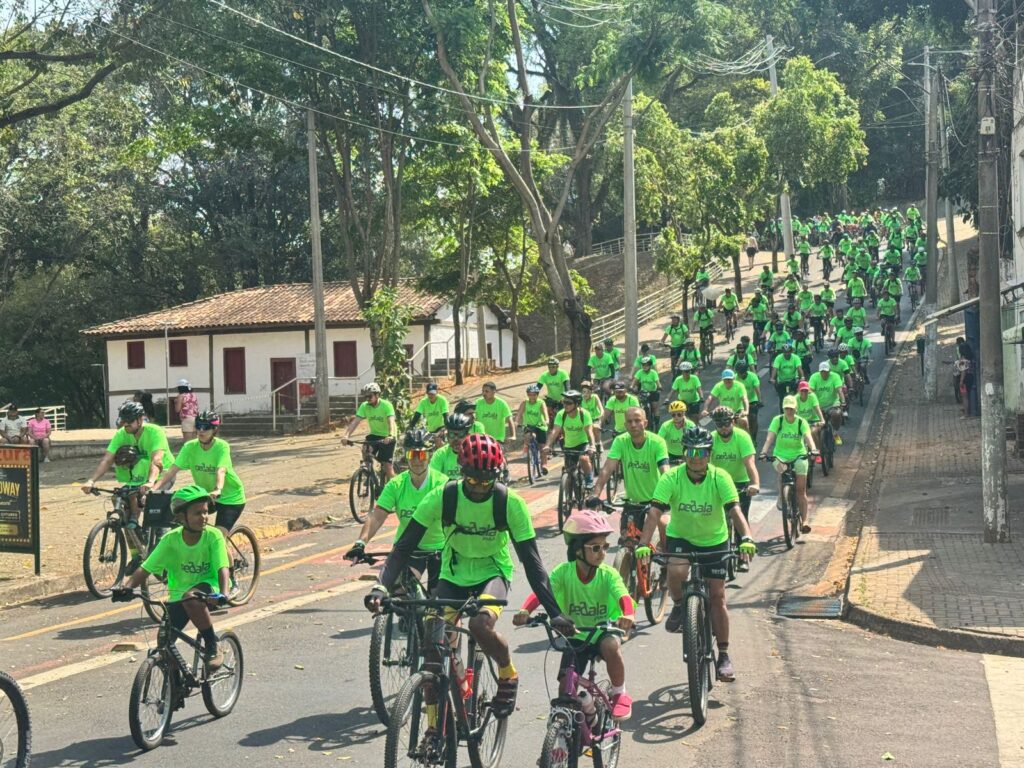 A imagem mostra um evento ciclístico de grande porte, com dezenas de pessoas pedalando por uma rua arborizada. Todos os participantes vestem camisetas verdes com o logo “Pedala”, indicando uma ação organizada — possivelmente uma campanha de incentivo ao ciclismo ou à mobilidade sustentável.