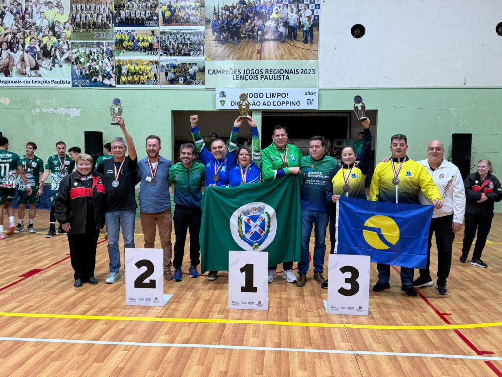 grupo de atletas e representantes posa em uma quadra esportiva durante cerimônia de premiação, exibindo troféus, medalhas e bandeiras ao lado do pódio de 1º, 2º e 3º lugares.