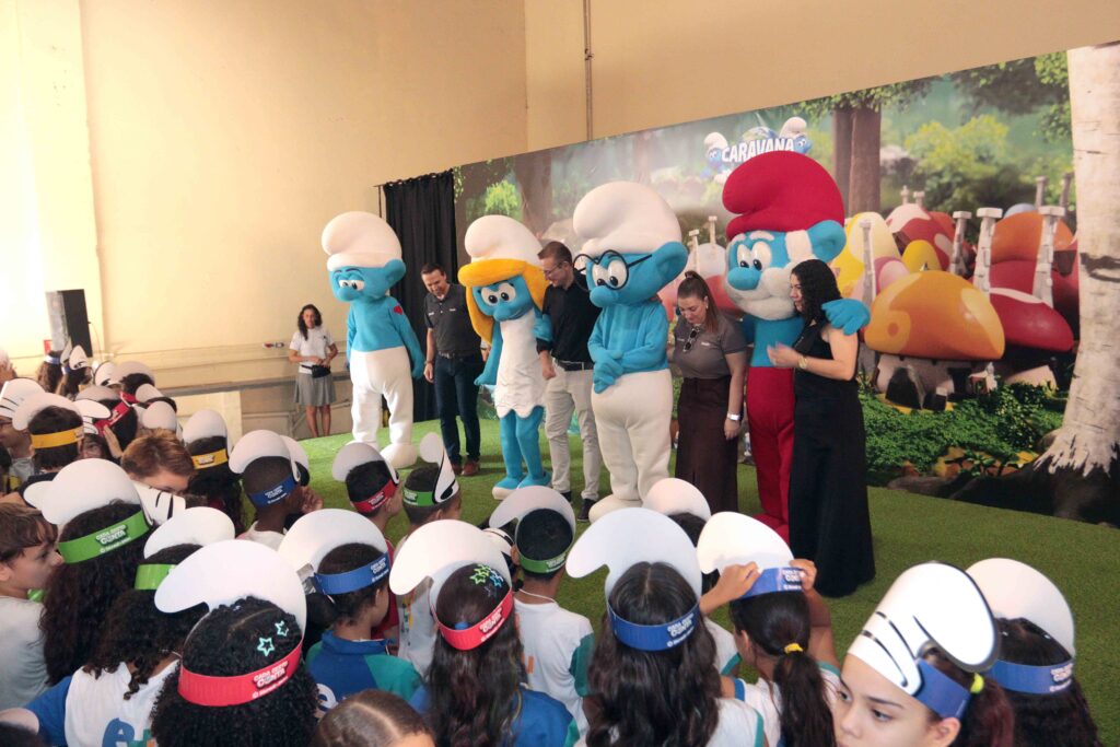 A imagem mostra um evento educativo em um espaço fechado, com um palco decorado com personagens dos Smurfs em tamanho gigante. Em frente ao palco, dezenas de estudantes usando viseiras temáticas assistem à apresentação. Também aparecem organizadores ao lado dos personagens, enquanto o público participa da atividade voltada à conscientização e ao aprendizado de forma lúdica.