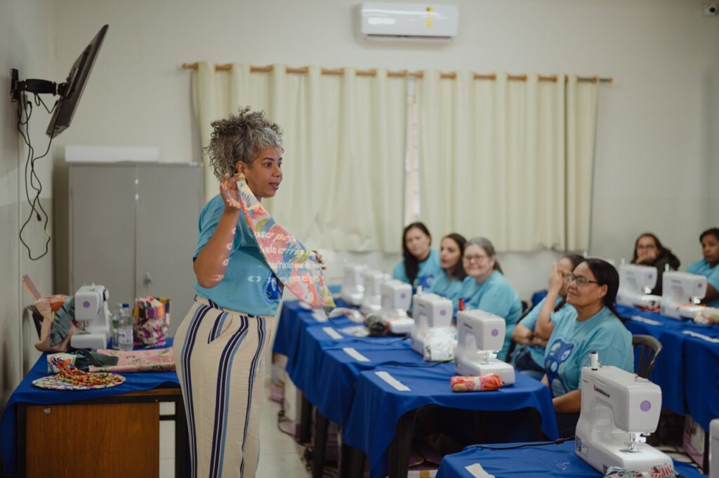 Uma sala de aula de costura com várias máquinas de costura sobre mesas cobertas por toalhas azuis. Na frente, uma pessoa apresenta um pedaço de tecido estampado para os participantes, que acompanham a demonstração sentados em seus lugares.