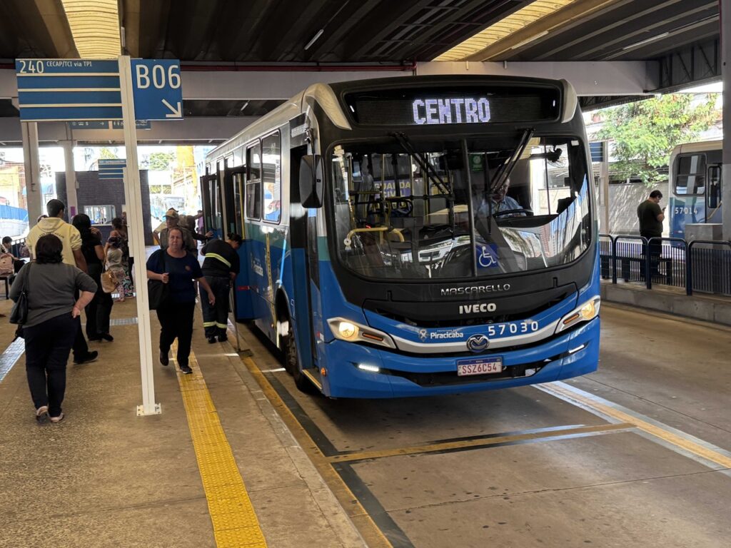 A imagem mostra um ônibus do transporte público urbano parado em uma plataforma de terminal, com o letreiro indicando o destino “Centro”. Passageiros circulam e aguardam o embarque ao lado do veículo, em uma área coberta e sinalizada para o atendimento das linhas municipais.