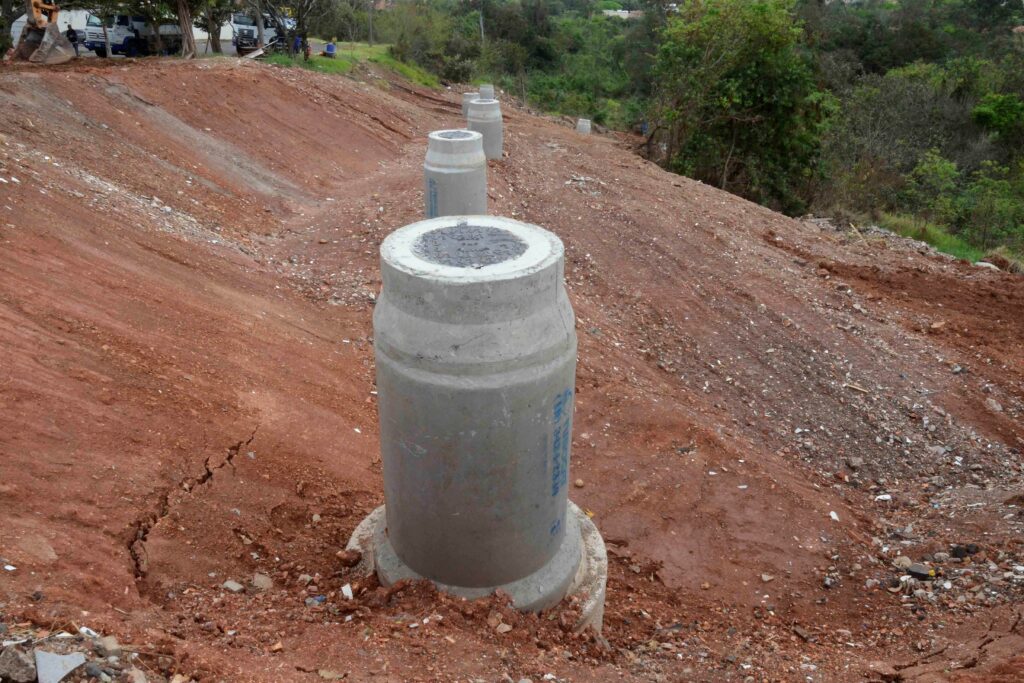 Os cilindros de concreto estão alinhados e fixados no solo inclinado. Eles fazem parte de um projeto de fundação ou estabilização de encosta. A cena mostra uma etapa inicial da engenharia civil em obras de infraestrutura.