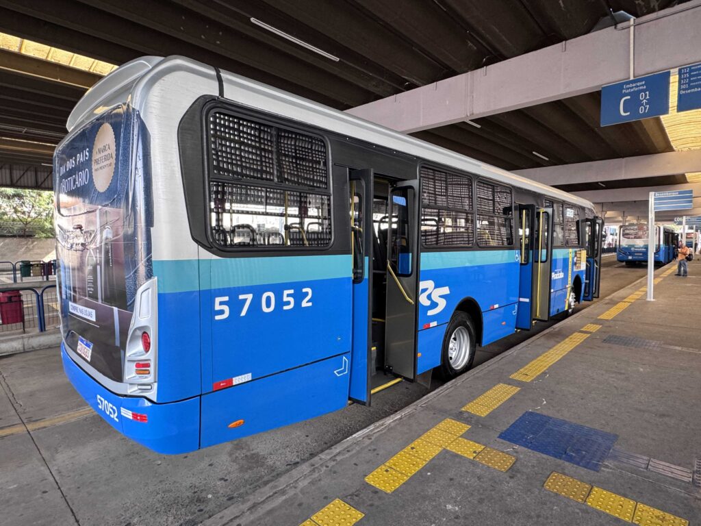 Um autocarro do transporte público nas cores azul, branca e cinzenta está parado na plataforma de um terminal de passageiros, com as portas abertas para embarque e desembarque.