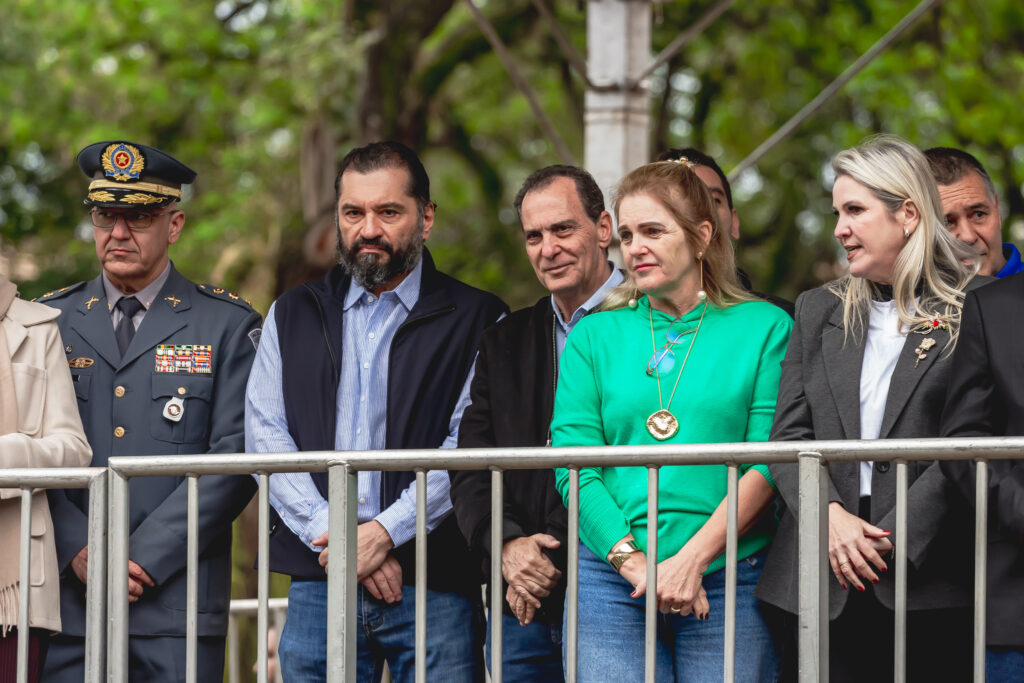vemos um grupo de pessoas reunidas atrás de uma grade metálica em um ambiente ao ar livre, com árvores ao fundo. Entre elas, destaca-se uma figura em uniforme militar adornado com medalhas e insígnias, sugerindo a presença de autoridade ou homenageado em um evento oficial. Os demais participantes estão em roupas civis, como casacos e suéteres, e mantêm postura atenta, reforçando o caráter solene da ocasião