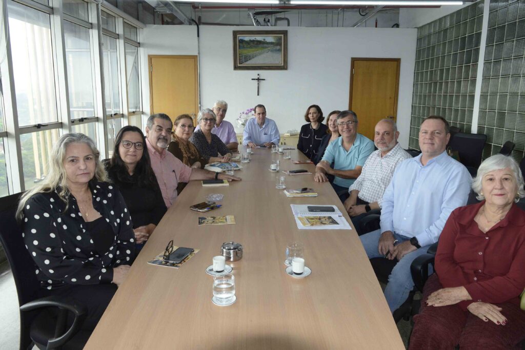 A imagem mostra um ambiente formal de reunião, com doze pessoas sentadas em torno de uma longa mesa de conferência. O espaço é iluminado por grandes janelas, e ao fundo há um quadro e um crucifixo, reforçando o caráter institucional do encontro. Sobre a mesa, vemos copos, garrafas de água, celulares, papéis e panfletos, elementos típicos de um cenário administrativo. A disposição dos participantes e os objetos sugerem uma discussão organizada, possivelmente ligada a decisões acadêmicas, empresariais ou comunitárias, destacando o aspecto colaborativo e profissional do momento.