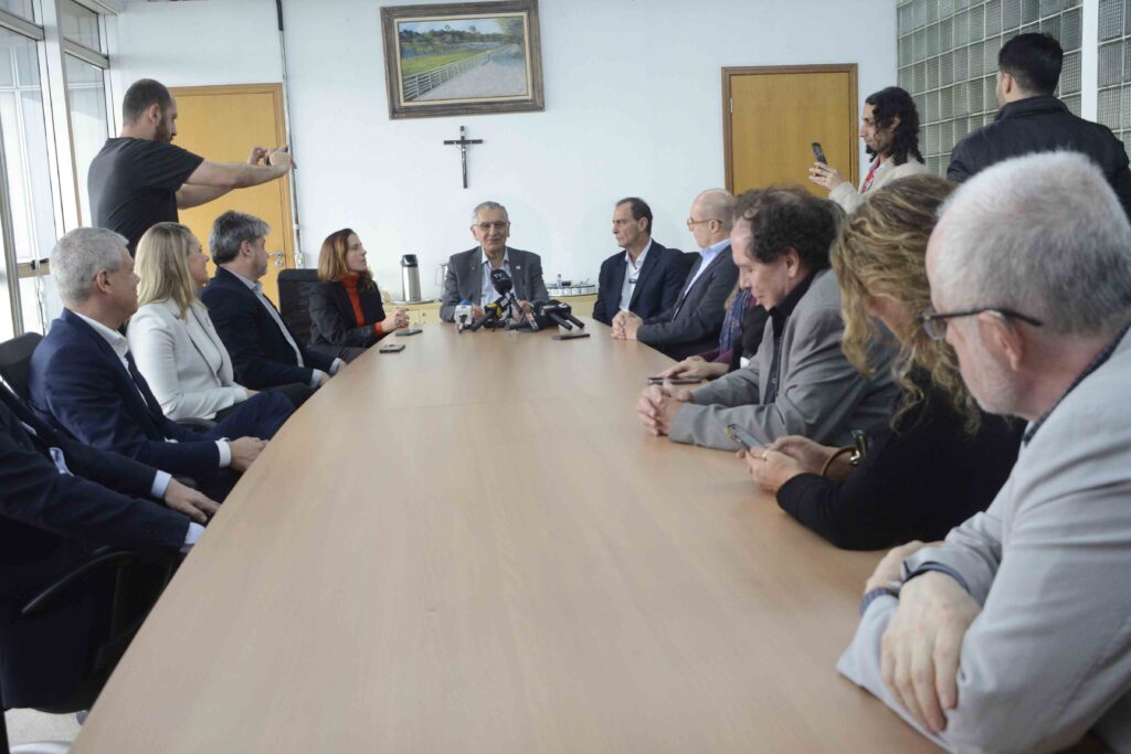 A imagem mostra uma reunião formal em uma sala simples, onde várias pessoas estão sentadas ao redor de uma grande mesa de conferência. Na cabeceira, alguém fala diante de microfones, sugerindo tratar-se de uma coletiva de imprensa ou discussão oficial. Os demais participantes acompanham atentamente, alguns com celulares em mãos, enquanto duas pessoas em pé registram o momento com fotos ou vídeos. O ambiente é marcado por paredes brancas, uma porta de madeira, um crucifixo e um quadro de paisagem, transmitindo sobriedade e foco no ato de comunicação e documentação.