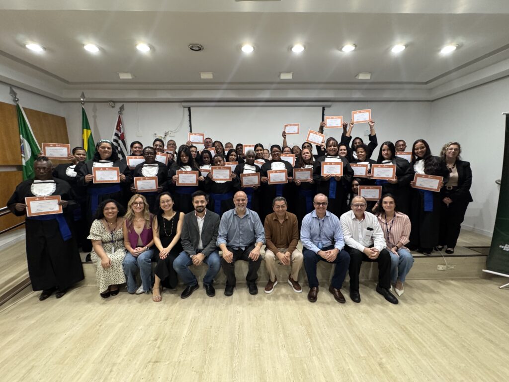 A imagem mostra uma grande cerimônia de formatura ou entrega de certificados em um auditório. Um grupo numeroso de pessoas está reunido em várias fileiras; muitas usam becas pretas com faixas azuis e seguram certificados com bordas laranjas, enquanto algumas os levantam para a foto. Na parte da frente, há pessoas sentadas usando roupas sociais e crachás. Ao fundo, aparecem uma tela de projeção, bandeiras — incluindo a do Brasil e a do estado de São Paulo —, iluminação embutida no teto e um piso claro de madeira.