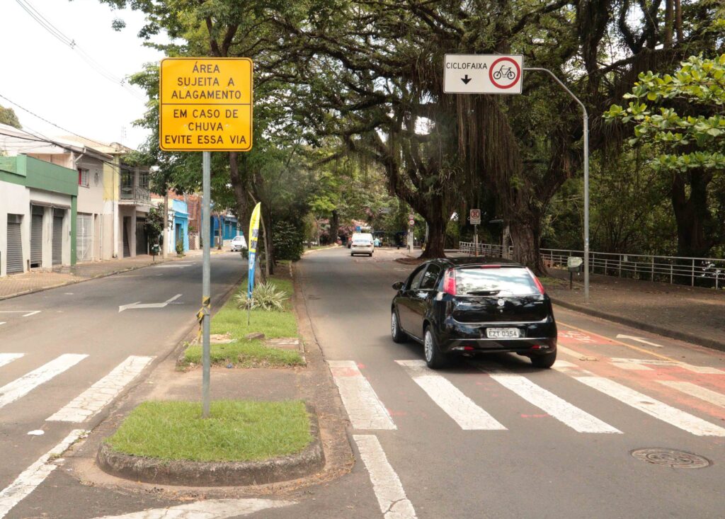 A imagem mostra uma via urbana arborizada com uma placa de advertência informando: “Área sujeita a alagamento. Em caso de chuva, evite essa via”.