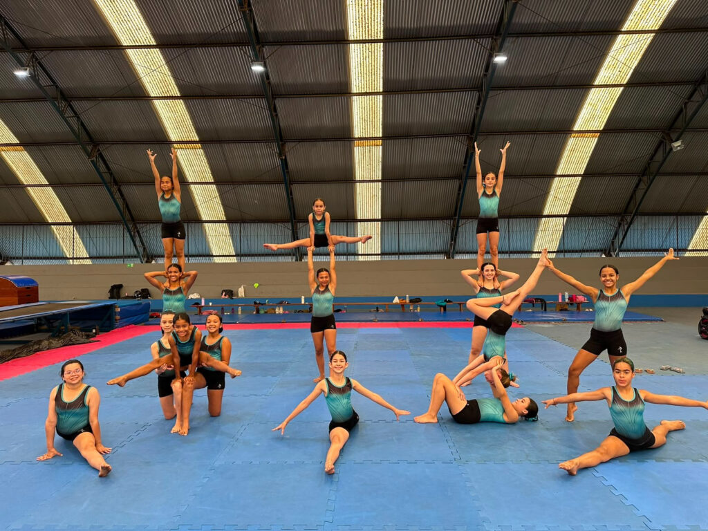 A imagem mostra um grupo de atletas em um ginásio realizando uma apresentação de ginástica/acrobacias em equipe.