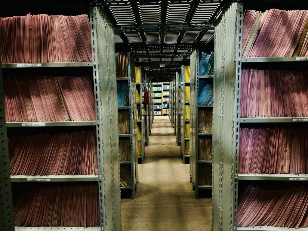 A imagem mostra um corredor de arquivo físico com estantes metálicas cheias de pastas coloridas — principalmente nas cores rosa, azul, amarela e bege — organizadas de forma sistemática. Cada prateleira possui etiquetas com numeração, indicando um método de catalogação ou classificação.