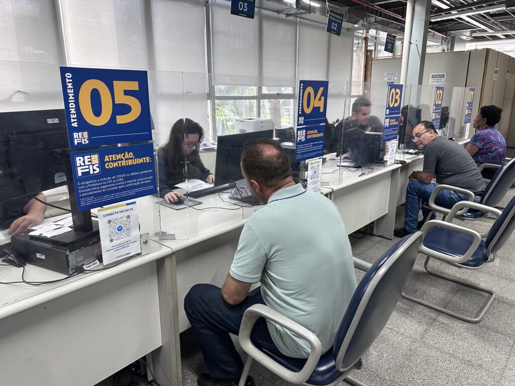 Guichês de atendimento ao público em uma repartição, com contribuintes sendo atendidos por servidores atrás de divisórias de proteção. Placas informam sobre o programa Refis 2026 e os números dos guichês de atendimento.