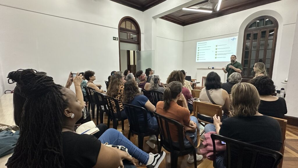 A imagem mostra uma palestra ou oficina sobre artesanato em uma sala ampla, com várias pessoas sentadas e atentas. Um homem está em pé, falando ao grupo, enquanto uma apresentação projetada na parede exibe o título “ARTESANATO” e o logotipo do Sebrae. O ambiente tem janelas e portas altas de madeira, teto com acabamento escuro, piso de madeira e cadeiras organizadas em fileiras. Algumas pessoas fazem anotações ou fotografam a apresentação.