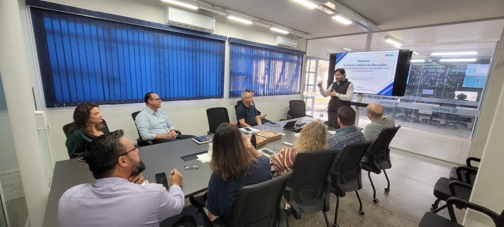 Participantes estão sentados em torno de uma mesa acompanhando uma apresentação. Um palestrante fala em frente a uma tela com o tema de controle interno, em um ambiente corporativo de treinamento.