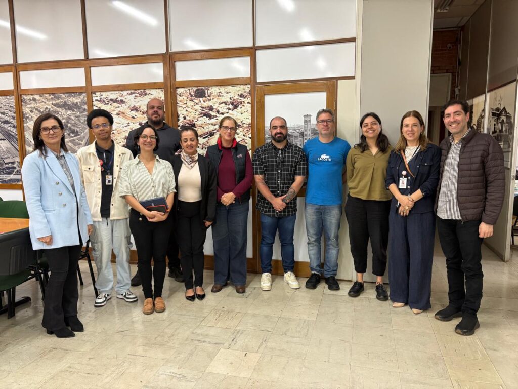 A imagem mostra um grupo de pessoas reunido em uma sala, participando de uma reunião ao redor de mesas, com um computador e uma tela exibindo informações. Ao fundo, há grandes imagens históricas de uma cidade.
