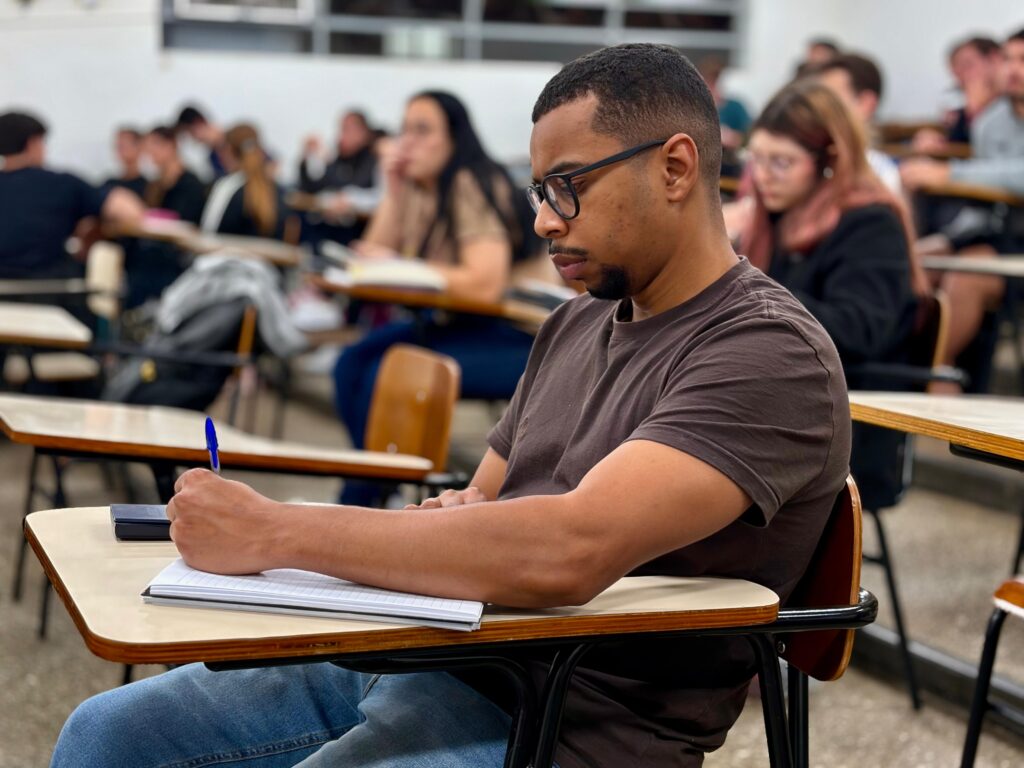Um estudante de óculos e t-shirt castanha concentrado a escrever num caderno durante uma aula numa sala cheia de alunos, representativo do ambiente do processo seletivo e da graduação na instituição.