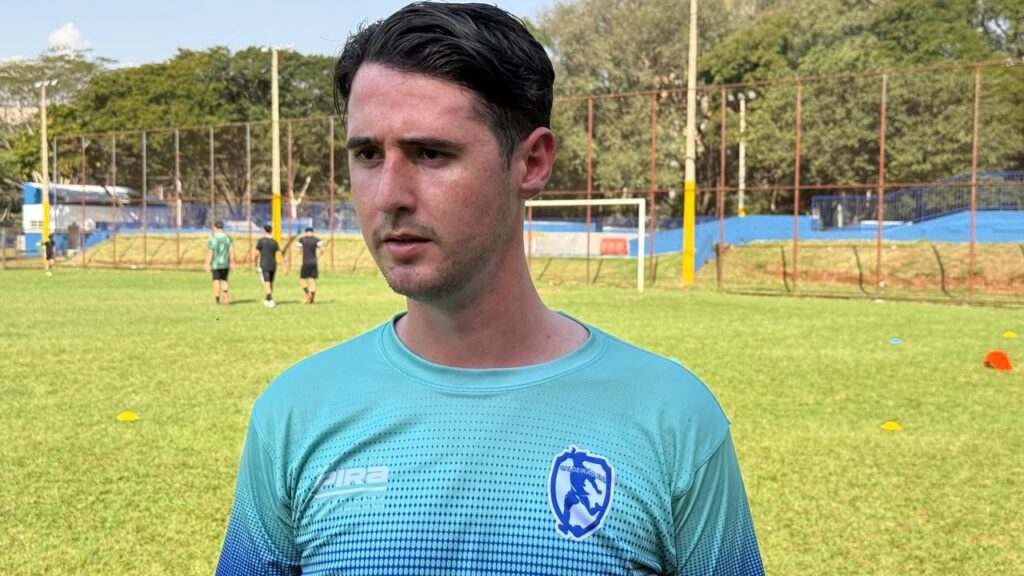A imagem mostra um homem jovem em primeiro plano, usando uma camisa esportiva azul-turquesa com um escudo no peito. Ele está em um campo de futebol gramado, olhando ligeiramente para o lado com uma expressão séria. Ao fundo, há outros jogadores treinando, uma trave, cones coloridos espalhados pelo campo, cercas altas e árvores. O dia está ensolarado.