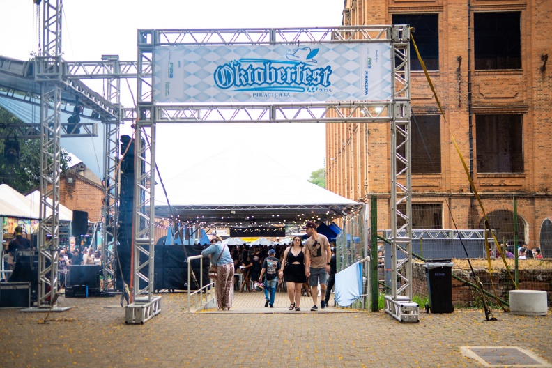 Um pórtico metálico de entrada com uma faixa do evento "Oktoberfest Piracicaba" dá acesso à área de festividades no Parque Engenho Central