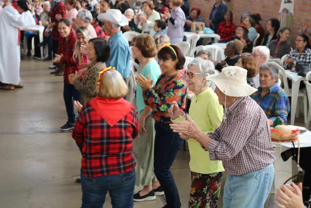 Participantes dançam e aplaudem em uma atividade coletiva, enquanto outras pessoas acompanham o evento sentadas ao redor do espaço.