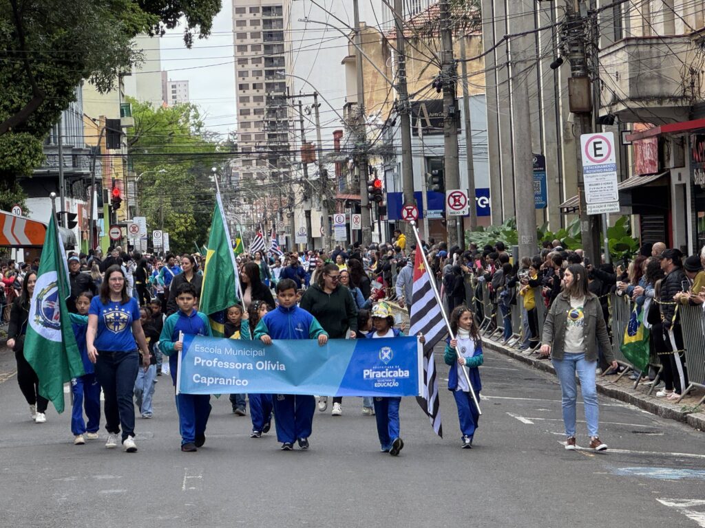 vemos um desfile escolar pelas ruas da cidade, com crianças e adultos marchando juntos e carregando bandeiras e faixas. O destaque é um grande banner escrito “Escola Municipal Professora Olívia Capranico – Piracicaba Educação”, que identifica a instituição participante. As crianças estão uniformizadas em azul e algumas seguram bandeiras, incluindo a do Brasil e a do estado de São Paulo. O cenário urbano, com prédios altos, fios elétricos e espectadores atrás das grades, reforça o clima de celebração cívica e comunitária, valorizando a educação e o orgulho local.