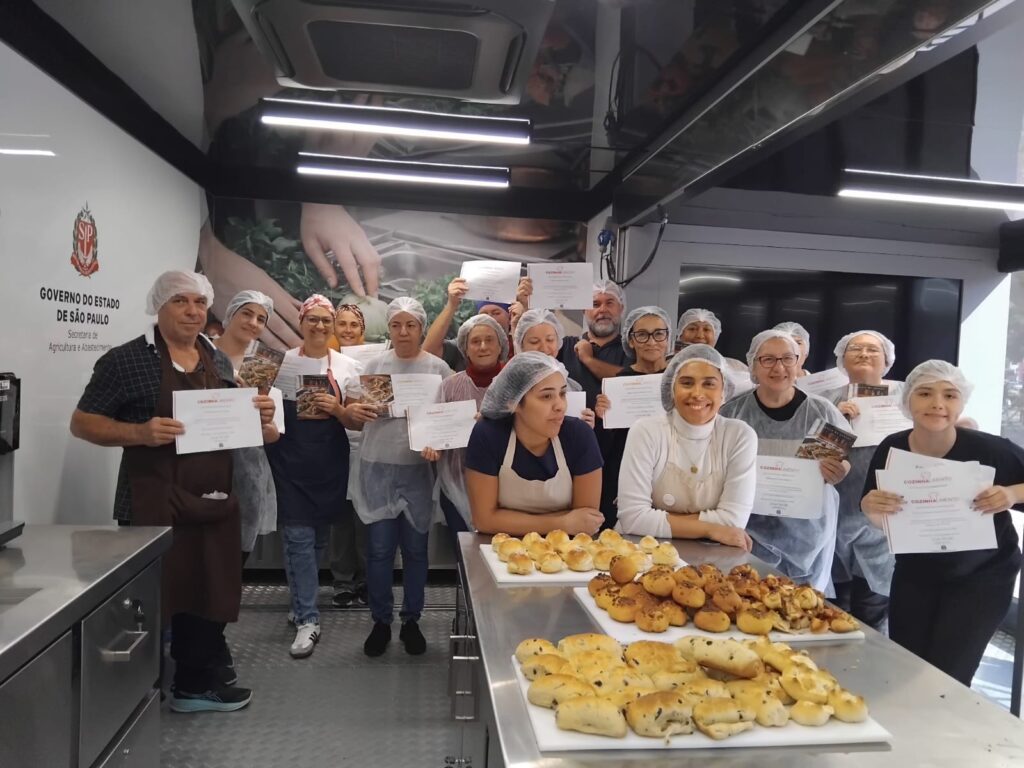 A imagem mostra um grupo de pessoas em uma aula de panificação comemorando a conclusão do curso. Todos estão usando toucas e aventais, e seguram certificados e apostilas, indicando que receberam formação prática.
