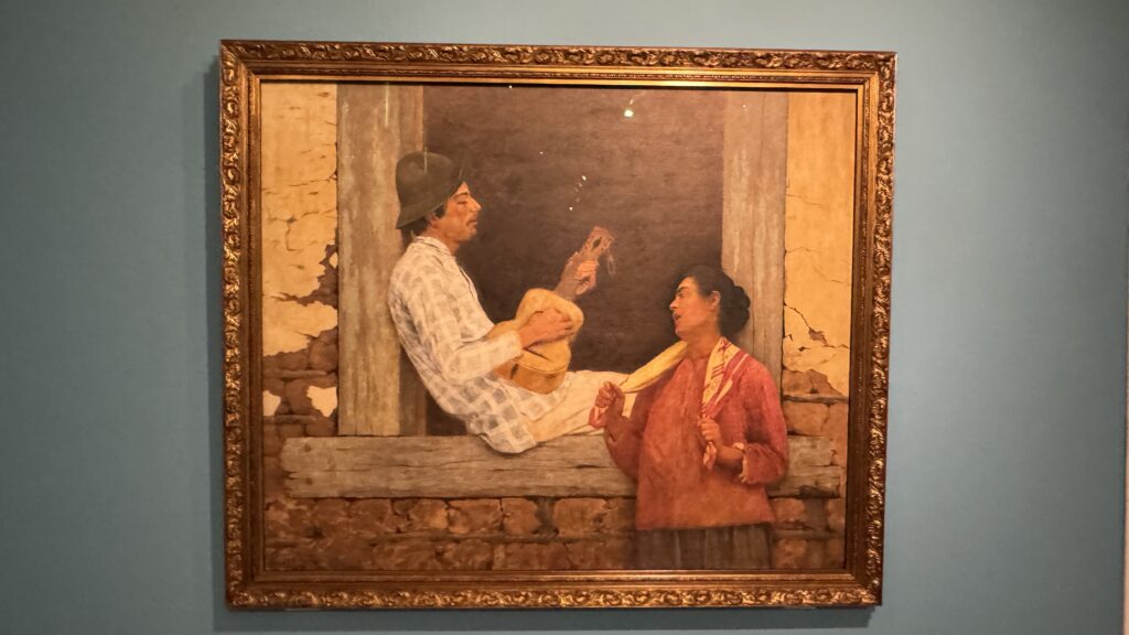 Quadro O Violeiro, de Almeida Júnior, retrata duas pessoas em cena rural diante de uma construção de alvenaria, em exposição na Pinacoteca.