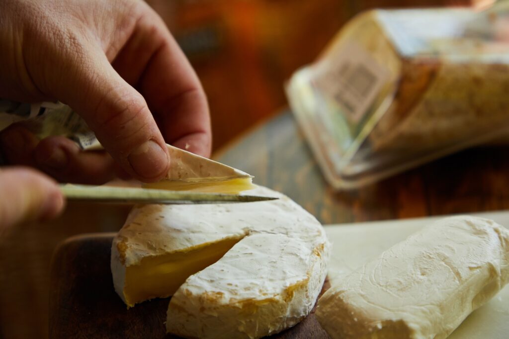 Esta fotografia em plano fechado captura um momento íntimo e artesanal, onde uma mão utiliza uma faca delicada para remover cuidadosamente a crosta de um queijo tipo Brie sobre uma tábua. A composição é focada na ação central, com a mão esquerda do indivíduo segurando firmemente a borda de uma fatia de queijo, enquanto a mão direita, visível apenas pela ponta da faca, realiza o corte preciso. O ângulo é ligeiramente elevado, permitindo uma visão clara da textura do queijo e da mecânica do gesto. A imagem utiliza uma profundidade de campo rasa, mantendo o queijo em foco nítido e desfocando suavemente tanto o primeiro plano quanto o fundo, o que direciona o olhar do espectador diretamente para a interação entre a lâmina e o alimento. O sujeito principal é um queijo Brie de formato circular, com sua casca branca aveludada e interior cremoso em tom amarelo-pálido, parcialmente cortado para revelar sua textura macia. Ao lado, encontra-se outro pedaço de queijo, possivelmente um queijo de cabra ou de pasta mole, com uma aparência mais lisa e uniforme. As mãos que manuseiam o queijo são práticas e estão posicionadas para exercer precisão, com o polegar exercendo pressão leve para segurar o alimento, enquanto a faca de lâmina fina e longa realiza o trabalho de descasque com controle e destreza. A imagem é uma fotografia digital realista que prioriza a fidelidade das texturas e a naturalidade do momento. A iluminação é suave e quente, sugerindo uma luz ambiente de cozinha ou área de preparação de alimentos, que realça os detalhes orgânicos da casca do queijo e as sombras sutis criadas pelo corte. Não há manipulações estilísticas evidentes; a técnica foca na clareza visual e na representação fiel da cena, utilizando um balanço de brancos que aquece os tons do queijo e da madeira da tábua, conferindo uma estética acolhedora e culinária. O cenário é uma cozinha ou espaço de preparação, onde a tábua de madeira escura serve de base para o queijo, conferindo um contraste rústico. Ao fundo, uma embalagem plástica de queijo aparece desfocada, indicando o ambiente doméstico de preparo. A iluminação lateral cria um jogo de luz e sombra que define os volumes e ressalta a cremosidade do produto. A atmosfera geral é de calma e atenção meticulosa aos detalhes, evocando uma sensação de apreciação pela culinária e pelo ritual de preparar ingredientes artesanais de qualidade para uma refeição especial.