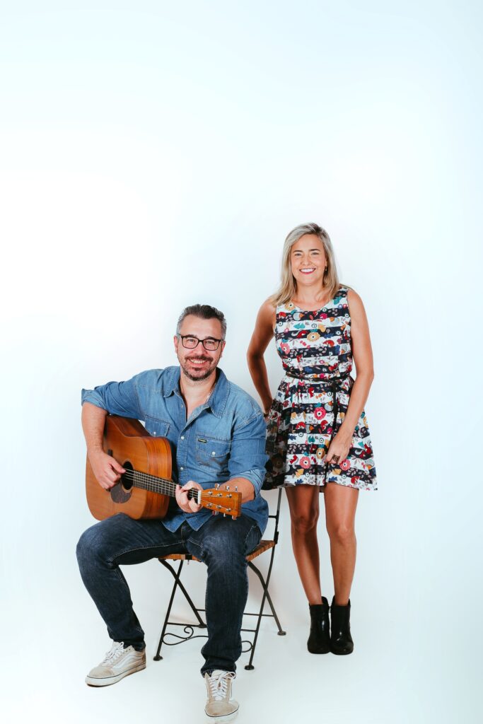 uma pessoa toca violão enquanto a outra posa ao lado, em um cenário de fundo branco, caracterizando uma apresentação acústica ou ensaio fotográfico de um projeto musical.