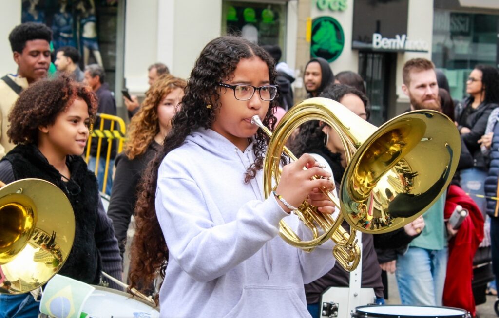 A imagem mostra uma apresentação musical em uma área urbana. Em primeiro plano, uma pessoa toca um instrumento de sopro de metal dourado, enquanto outras pessoas com instrumentos da banda aparecem ao redor.