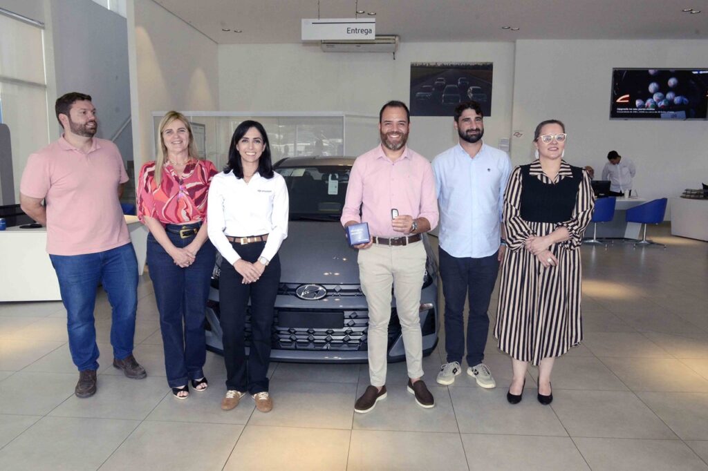 A imagem mostra seis pessoas sorridentes em uma concessionária Hyundai, posicionadas em frente a um carro cinza. O homem ao centro segura uma chave e uma caixa azul com a inscrição “Momento especial”, indicando a entrega de um veículo novo. Acima deles há uma placa com a palavra “Entrega”, reforçando o contexto de celebração. O ambiente é moderno e bem iluminado, com mesas e cadeiras ao fundo, típicas de um showroom.
