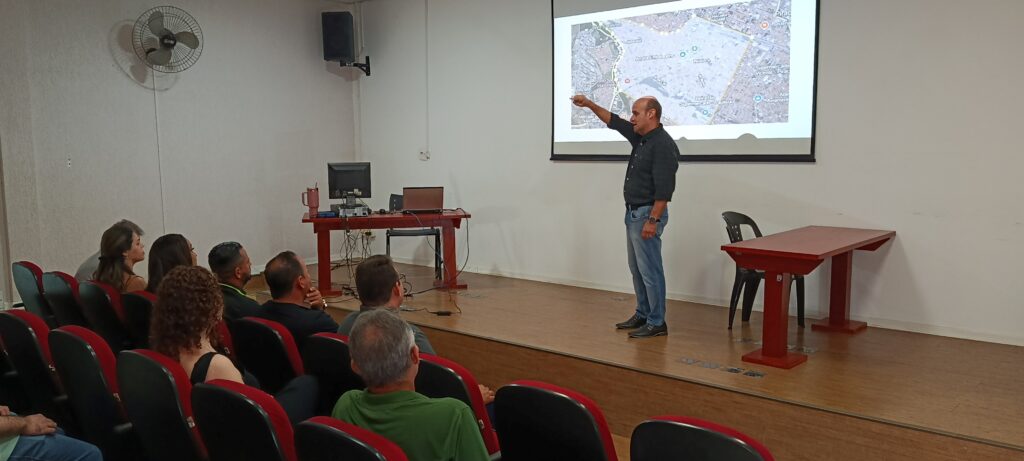 Um palestrante de pé, em cima do palco, aponta para um mapa projetado numa tela ao fundo enquanto apresenta o planeamento da ação aos presentes no auditório.
