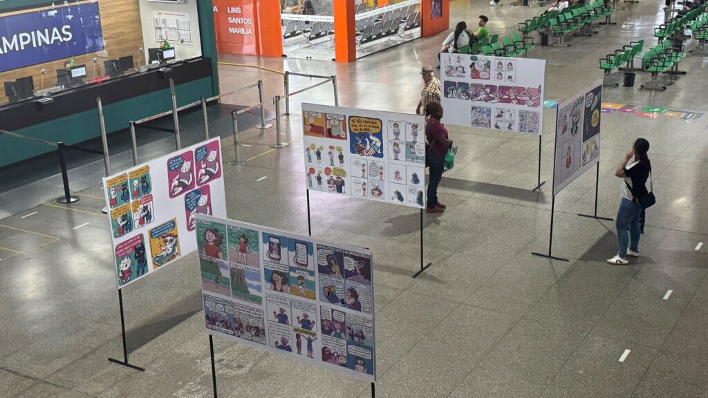 Mostra de tirinhas e histórias em quadrinhos expostas em painéis dentro de um terminal rodoviário, observadas por visitantes.