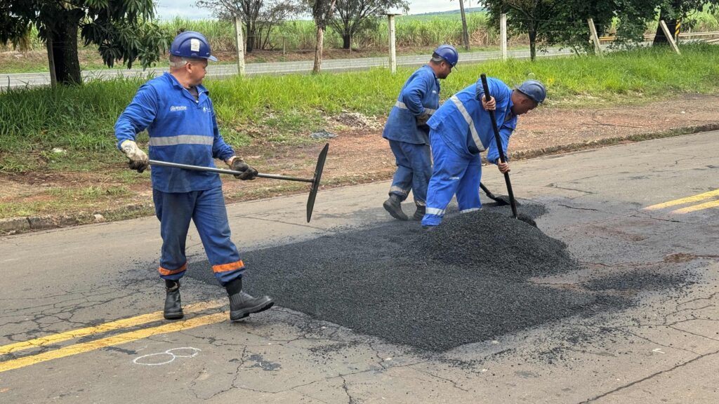 A imagem mostra três trabalhadores realizando manutenção em uma estrada, todos vestidos com uniformes azuis com faixas refletivas e capacetes de segurança. Eles estão espalhando e nivelando massa asfáltica sobre uma área danificada do pavimento, utilizando pás e enxadas.