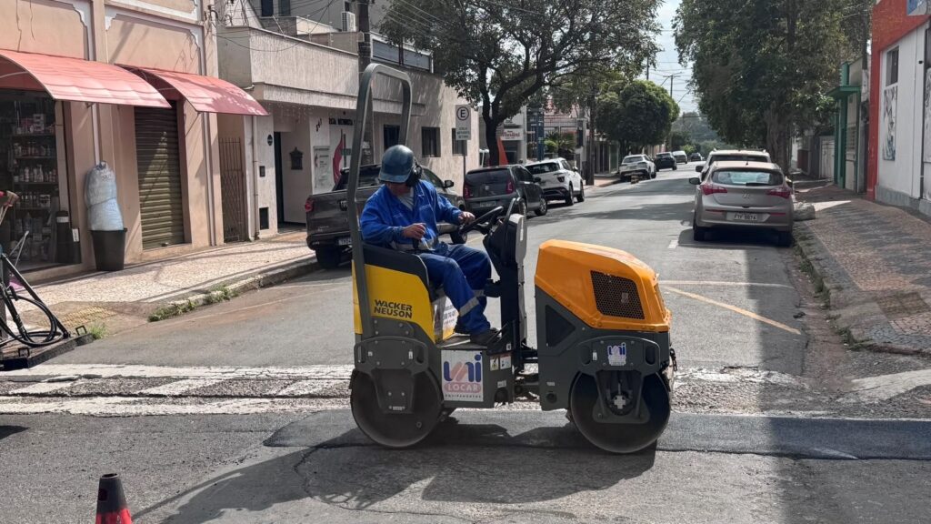 A imagem mostra uma rua residencial e comercial durante uma obra de pavimentação. No centro, um trabalhador usando uniforme azul e capacete opera um rolo compactador pequeno, amarelo e preto, com a identificação “Wacker Neuson” e “Uni Locar”. Há um cone de sinalização no primeiro plano, uma faixa de asfalto recém-aplicado e várias áreas remendadas na rua. Ao fundo, aparecem carros estacionados, árvores, calçadas de pedras portuguesas, fachadas de lojas e muitos fios elétricos. Há também uma placa indicando restrição de estacionamento.