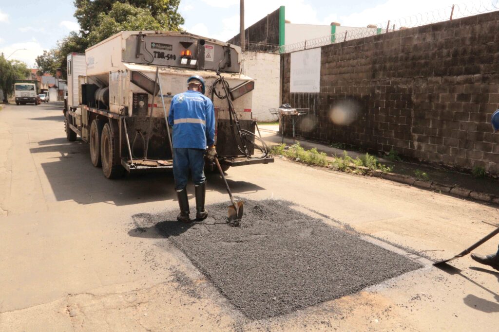 A imagem mostra uma operação de tapa-buraco em uma rua. Um caminhão de manutenção viária está parado enquanto trabalhadores utilizam pás para espalhar asfalto sobre um trecho da pista. Ao fundo, há muros, árvores e outros veículos, em um cenário urbano durante o dia.